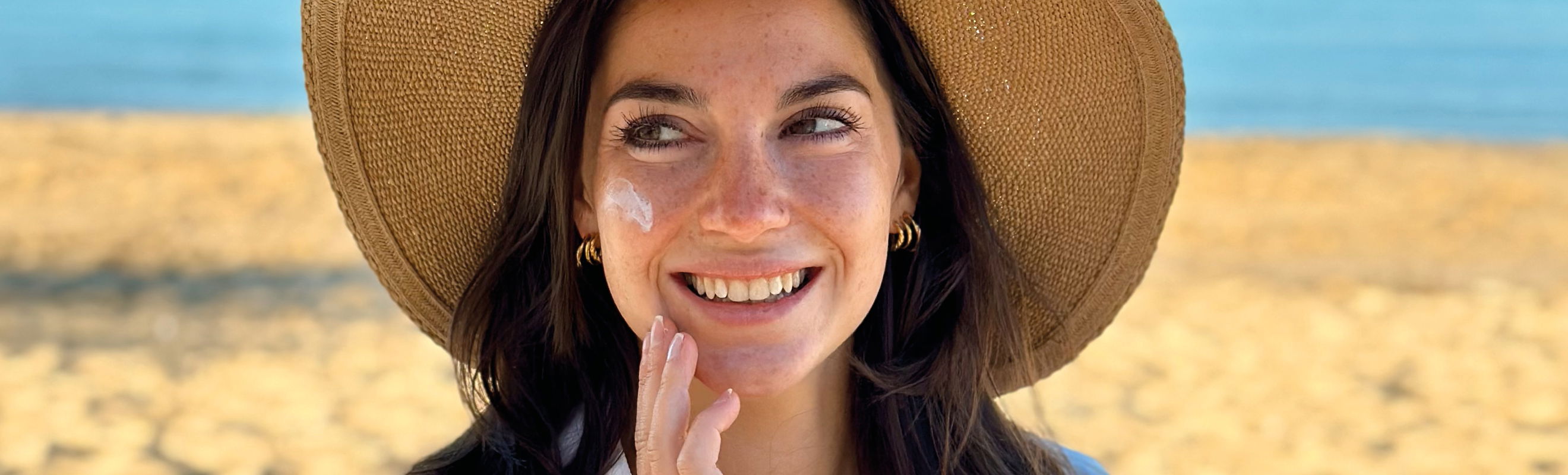 Brünette Frau mit Sonnenhut cremt am Strand ihr Gesicht ein