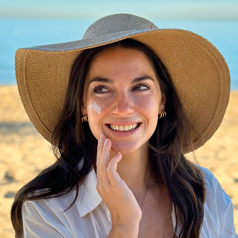 Brünette Frau mit Sonnenhut cremt am Strand ihr Gesicht ein
